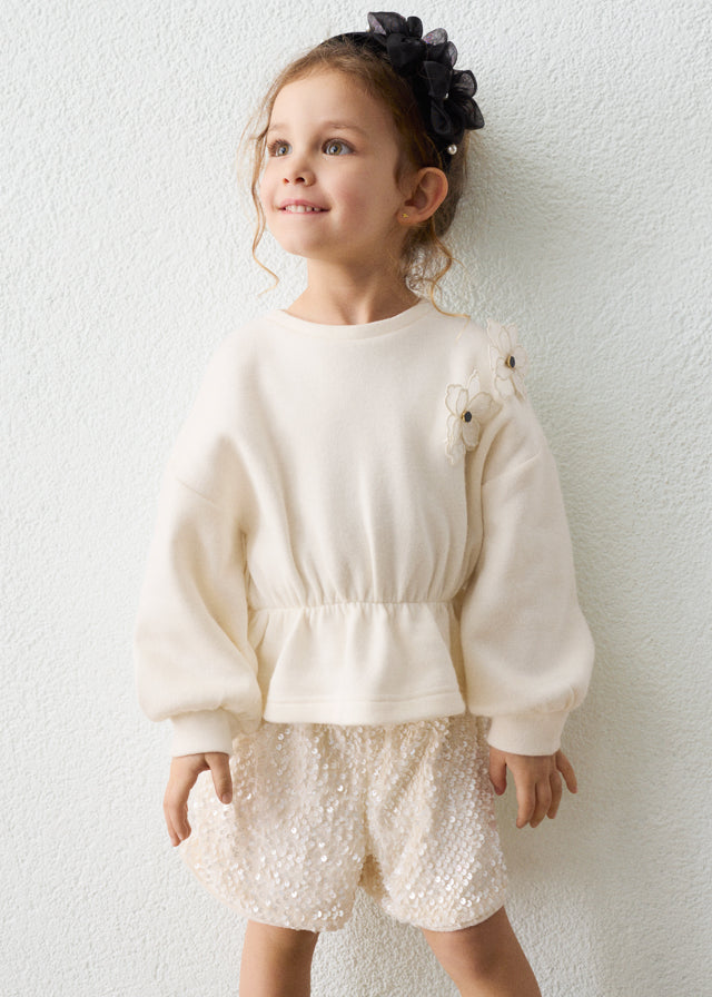 Young girl wearing a cream sweater and white shorts with a textured background
